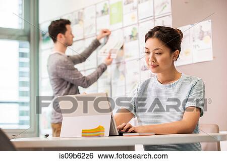 Female digital designer typing on laptop and colleague arranging mood board in office View Large Photo Image Stock Photograph - Female digital designer typing on laptop and colleague arranging mood board in office. Fotosearch