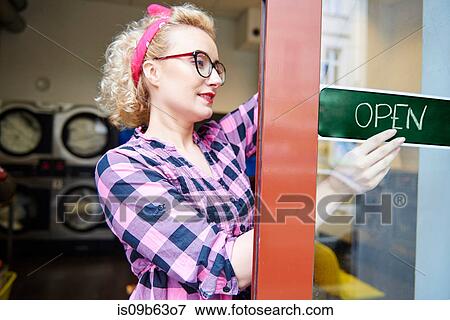 Stock Photo - Female small business owner putting up open sign on laundrette door. Fotosearch