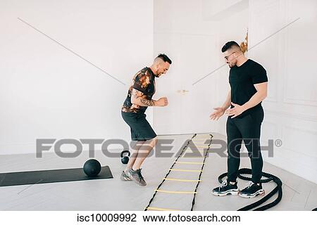 Stock Image - Fitness instructor observing man using agility ladder in studio. Fotosearch