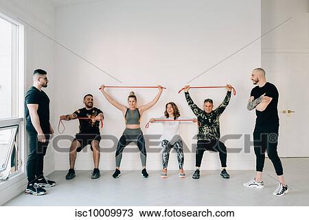 Stock Image - Fitness instructors observing friends doing squat against wall with resistance band in studio. Fotosearch