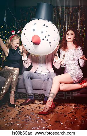 Stock Image - Friends sitting together at party, man holding snowman head in front of face, women laughing. Fotosearch