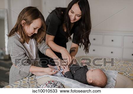 Stock Image - Girl helping mother change baby brother's diaper on bed. Fotosearch