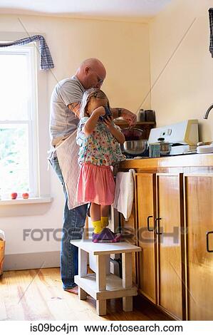 Picture - Girl on stool drinking while father prepares food in kitchen. Fotosearch