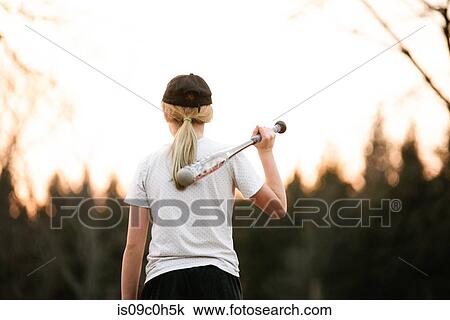 Stock Image - Girl with baseball bat over her shoulder in rural field, rear view. Fotosearch