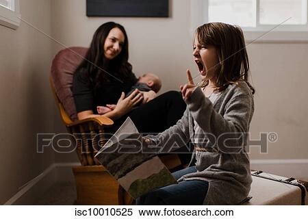Stock Photography - Girl with book pulling faces, while mother cradles baby brother in living room armchair. Fotosearch