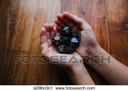 Stock Image - Girl with cupped hands at table holding role playing dice, close up. Fotosearch
