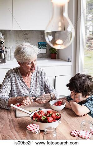 Stock Image - Grandmother sitting at kitchen table, preparing strawberries, grandson sitting beside her, watching. Fotosearch