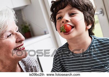 Stock Image - Grandson eating strawberry, grandmother sitting beside him, smiling. Fotosearch