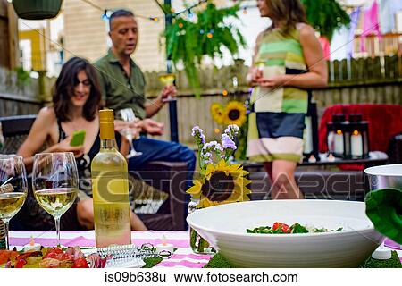 Stock Photograph - Group of people at garden party, holding wine glasses, bottle of wine and food on serving dishes in foreground. Fotosearch