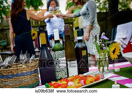 Stock Image - Group of women at garden party, holding wine glasses, making toast, wine bottles on table in foreground. Fotosearch