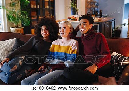 Stock Image - Happy mid adult man and young female friends watching tv on living room sofa. Fotosearch