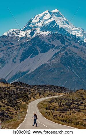 Stock Image - Hiker crossing road leading towards mountains, Wanaka, Taranaki, New Zealand. Fotosearch