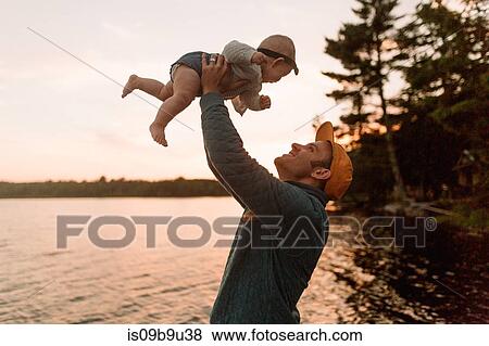 Stock Photo - Man holding up baby daughter at lakeside. Fotosearch