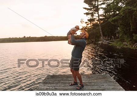 Stock Image - Man holding up baby daughter on lake pier. Fotosearch