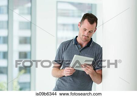 Man leaning against wall, looking at digital tablet View Large Photo Image Picture - Man leaning against wall, looking at digital tablet. Fotosearch