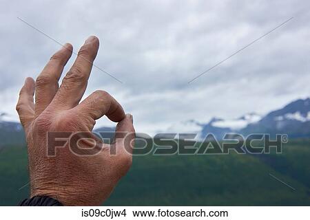 Picture - Man making OK sign in mountain landscape, close up of hand, Valdez, Alaska, USA. Fotosearch
