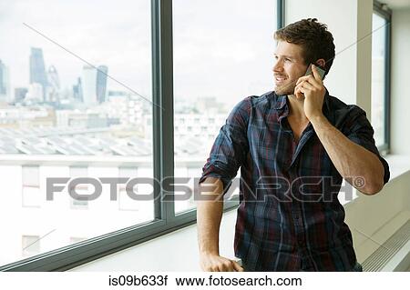 Stock Image - Man standing beside window in empty office space, using smartphone. Fotosearch