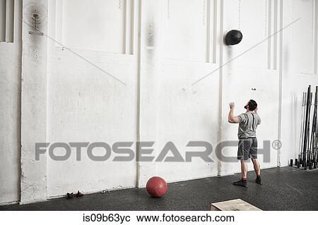 Stock Image - Man throwing fitness ball against wall in cross training gym. Fotosearch