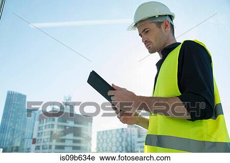 Stock Image - Man wearing hard hat and hi vis vest, looking at digital tablet. Fotosearch