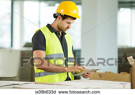 Stock Image - Man wearing hi vis vest, standing in newly constructed space, looking at digital tablet. Fotosearch