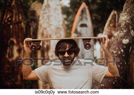Stock Photo - Mid adult male skateboarder carrying skateboard on head by old surfboard structure in park, portrait, Haiku, Hawaii, USA. Fotosearch