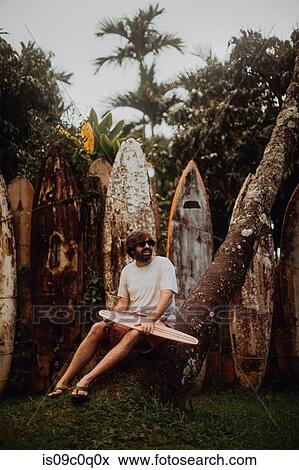 Stock Photo - Mid adult male skateboarder sitting on tree trunk by old surfboard structure in park, Haiku, Hawaii, USA. Fotosearch