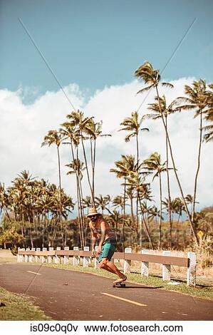 Stock Photo - Mid adult male skateboarder wearing straw hat skateboarding on rural road, Haiku, Hawaii, USA. Fotosearch