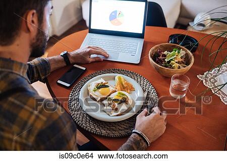 Picture - Mid adult man about to eat fried egg meal at table, looking at laptop, over shoulder view. Fotosearch