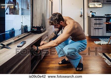 Stock Image - Mid adult man crouching to open living room cupboard. Fotosearch