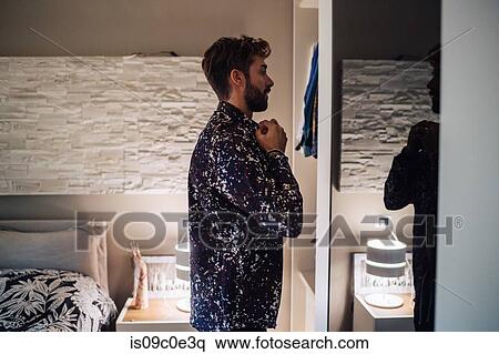 Stock Photography - Mid adult man fastening shirt buttons at bedroom mirror. Fotosearch