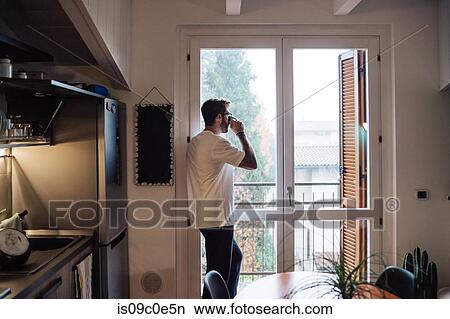 Picture - Mid adult man holding drinking coffee and looking out through apartment window. Fotosearch