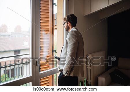 Mid adult man holding smartphone gazing out through apartment window View Large Photo Image Picture - Mid adult man holding smartphone gazing out through apartment window. Fotosearch