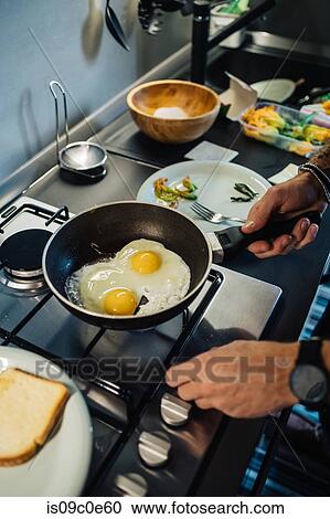 Stock Image - Mid adult man preparing fried eggs at kitchen hob, cropped close up. Fotosearch