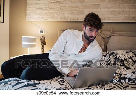 Picture - Mid adult man reclining on bed looking at laptop. Fotosearch