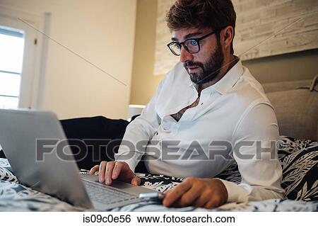 Stock Photograph - Mid adult man reclining on bed looking at laptop. Fotosearch