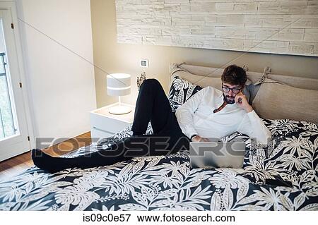 Stock Photo - Mid adult man reclining on bed using laptop. Fotosearch