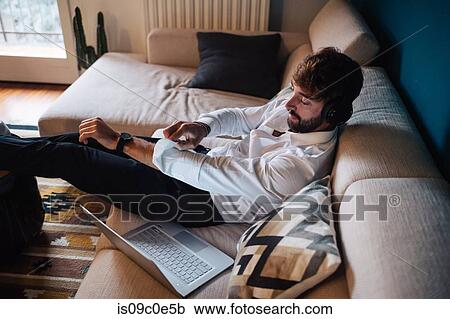 Stock Image - Mid adult man reclining on sofa listening to headphones while rolling up sleeves. Fotosearch