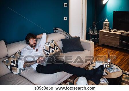 Stock Image - Mid adult man reclining on sofa listening to headphones and typing on laptop. Fotosearch