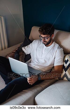 Picture - Mid adult man reclining on sofa using laptop. Fotosearch