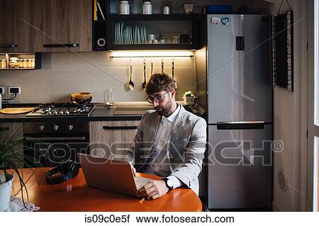 Stock Image - Mid adult man sitting at kitchen table typing on laptop. Fotosearch