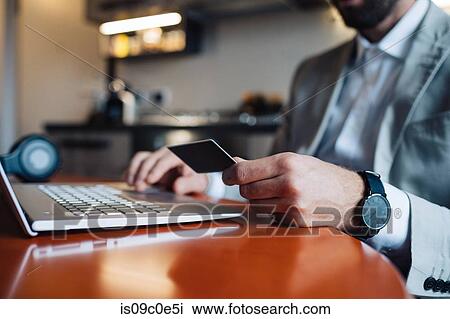 Mid adult man sitting at kitchen table with laptop and credit card, cropped View Large Photo Image Stock Image - Mid adult man sitting at kitchen table with laptop and credit card, cropped. Fotosearch