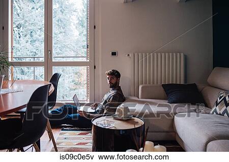 Stock Photo - Mid adult man sitting on living room floor using laptop, portrait. Fotosearch