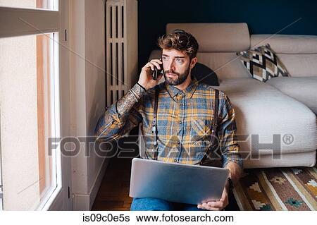 Mid adult man sitting on living room floor with laptop making smartphone call View Large Photo Image Stock Photograph - Mid adult man sitting on living room floor with laptop making smartphone call. Fotosearch