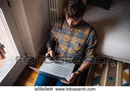Stock Photograph - Mid adult man sitting on living room floor with laptop using smartphone. Fotosearch