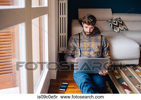 Stock Photography - Mid adult man sitting on living room floor looking at laptop. Fotosearch