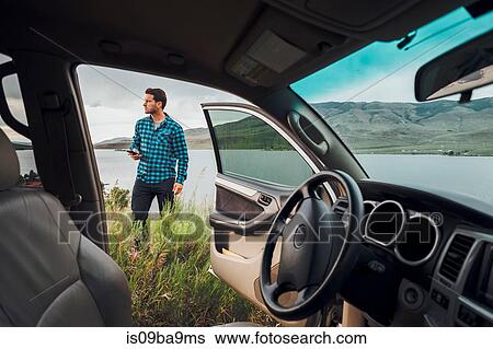 Stock Photograph - Mid adult man standing beside Dillon Reservoir, holding smartphone, view through parked car, Silverthorne, Colorado, USA. Fotosearch