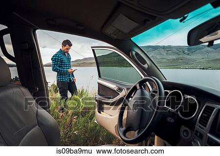 Stock Photograph - Mid adult man standing beside Dillon Reservoir, holding smartphone, view through parked car, Silverthorne, Colorado, USA. Fotosearch