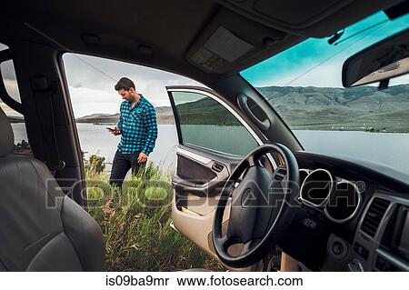 Stock Photography - Mid adult man standing beside Dillon Reservoir, holding smartphone, view through parked car, Silverthorne, Colorado, USA. Fotosearch