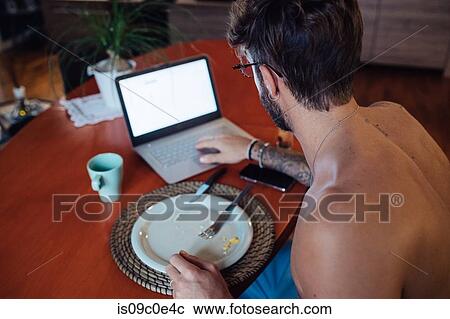 Stock Image - Mid adult man typing on laptop at breakfast table. Fotosearch