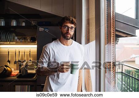 Stock Photo - Mid adult man with coffee by apartment window, portrait. Fotosearch